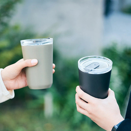 Two hands holding insulated travel mugs outdoors with a blurred green background