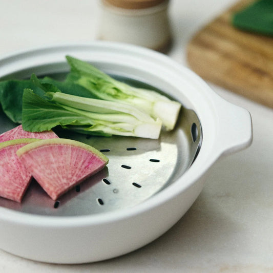 White ceramic steamer basket with sliced vegetables on a light background