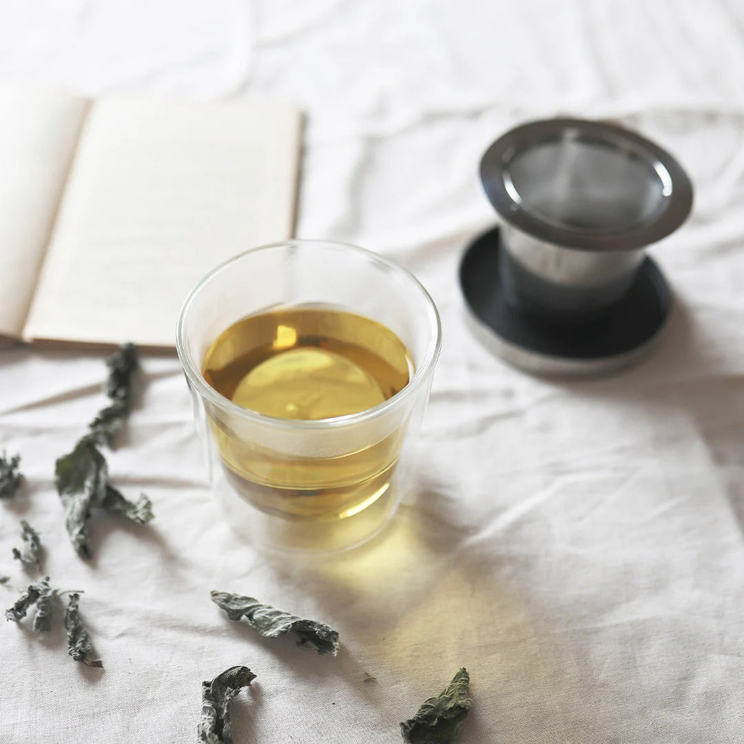 Glass of green tea with tea leaves and a teapot on a light surface