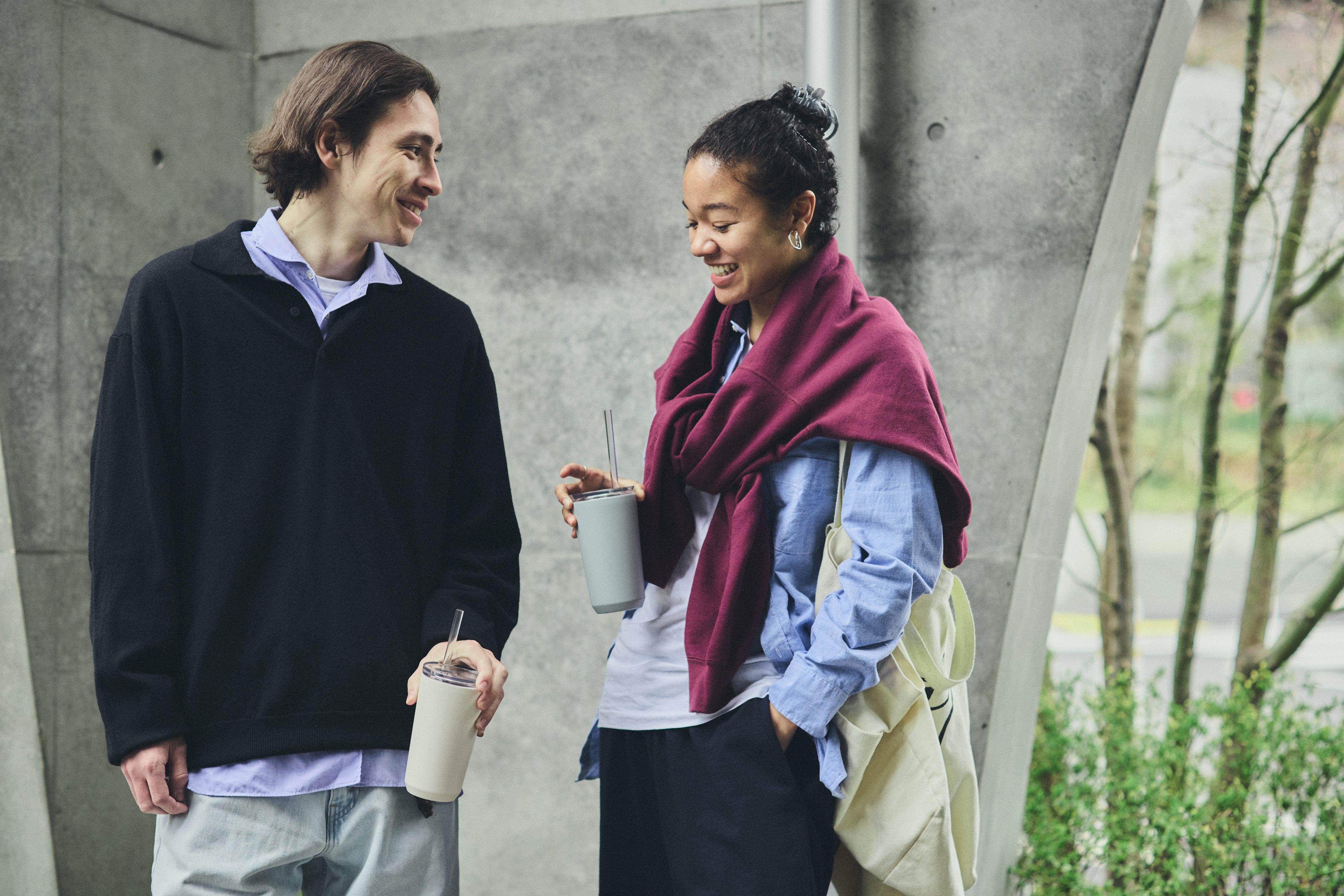 Two people standing outdoors, holding drinks, with a concrete wall and greenery in the background.